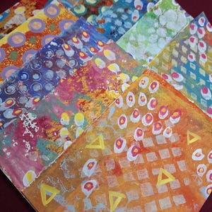 May include: A collection of rectangular cards featuring abstract painted designs in vibrant colors including orange, yellow, pink, blue, and green. The surfaces display layered patterns of circles, grids, and triangles created with acrylic paint.