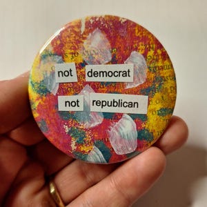 May include: A round button with a colorful abstract background of red, yellow, and blue. The button has the words "not democrat" and "not republican" printed on it.