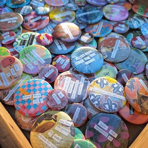 Puede incluir: Una colección de botones redondos de colores con varios mensajes inspiradores y obras de arte. Los botones están dispuestos en una bandeja de madera.