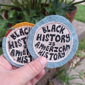 May include: Two round fabric patches with the text "BLACK HISTORY IS AMERICAN HISTORY" in black lettering. One patch has a light blue border, the other has a yellow and red border. The patches are held in a hand.