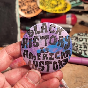 Puede incluir: Un botón redondo con el texto "BLACK HISTORY IS AMERICAN HISTORY" en letras negras sobre un fondo morado y azul. Otros botones con texto similar son visibles en el fondo.