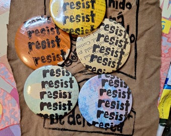 May include: Four round buttons with the word "resist" printed on them in black lettering. The buttons are made of different materials and have different colors and patterns. The buttons are arranged in a circle on a brown fabric background.