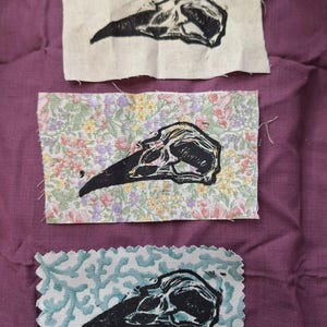 May include: Three fabric patches with black bird skull prints. The top patch is plain, the middle has a floral pattern, and the bottom has a teal and white design. The prints are detailed and the fabric is rectangular.