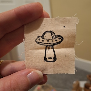 May include: A small, beige fabric patch featuring a black illustration of a UFO. The UFO design includes a saucer-shaped top with portholes and a rectangular base. The edges of the patch are frayed.