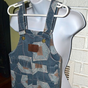 May include: Blue denim overalls with a geometric pattern of brown and white squares. The overalls have adjustable straps with brass buckles.