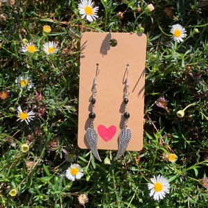 Puede incluir: Un par de pendientes de plata con cuentas negras y alas de ángel plateadas. Los pendientes se muestran en una tarjeta marrón con un corazón rojo.