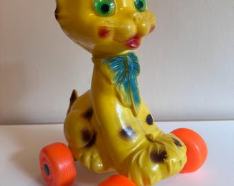 Könnte beinhalten: Ein gelbes Katzenspielzeug aus Kunststoff auf vier leuchtend orangefarbenen Rädern. Die Katze hat große grüne Augen, dunkelbraune Flecken und eine blaue Schleife um den Hals. Sie hat einen lächelnden Ausdruck mit roten Wangen und Lippen sowie einen spitzen Schwanz.