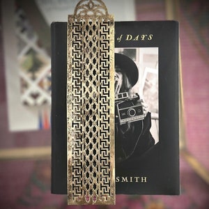 May include: A gold-colored metal bookmark with a geometric design is placed on a book. The bookmark has an ornate top and a rectangular shape with a repeating pattern. The book has a black cover with the title "of DAYS" and the author's name "SMITH".