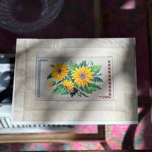 May include: A vintage needlepoint floral design featuring yellow and purple flowers with green leaves. The artwork is framed with blue and red stitched borders. The piece is displayed on a glass table.