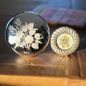 May include: Two decorative round ornaments. One is a clear glass ball with a white floral design. The other is a silver metal ornament with a floral design on the inside.