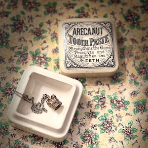 May include: A vintage-style box of "ARECANUT TOOTH PASTE" with ornate lettering, alongside a small white dish holding a silver crown pendant necklace. The background is a floral pattern.