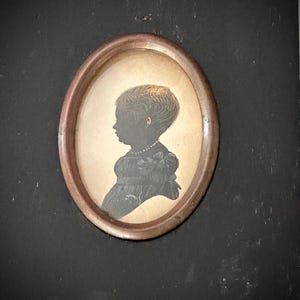 May include: A black and white silhouette of a person with short hair, wearing a collared shirt and a bow tie, framed in a black oval frame with a copper-colored border.
