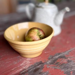 May include: A yellow ceramic bowl with two horizontal white stripes around the exterior, containing a single green and red apple. Behind the bowl sits a speckled white ceramic teapot with a gold-colored knob on the lid.