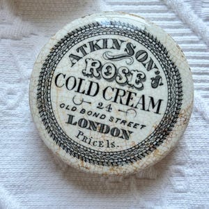 May include: A round, antique Atkinson's Rose Cold Cream container. The cream jar has a white background with black text and a decorative border. The text includes "24 Old Bond Street London". The container has a cracked, aged appearance.
