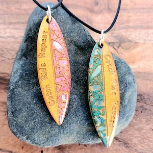 May include: Two wooden surfboard-shaped necklaces with the text "Catch Ride Repeat" on each. The necklaces are on a gray rock and have a black cord.