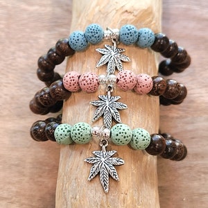 May include: Three bracelets with a cannabis leaf charm. Each bracelet features a combination of dark brown wooden beads and colorful lava stone beads in blue, pink, and green. The charms are silver-colored.