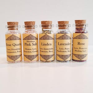 May include: Five small glass bottles with cork tops, each filled with different herbs and crystals. The bottles are labeled with the names of the herbs and crystals, including Rose Quartz, Himalayan Pink Salt, Linden, Lavender, and Rose. Each label also includes a description of the herb or crystal's properties, such as love, friendship, compassion, peace, cleanse, protection, growth, immortality, sleep, purification, healing, and luck.