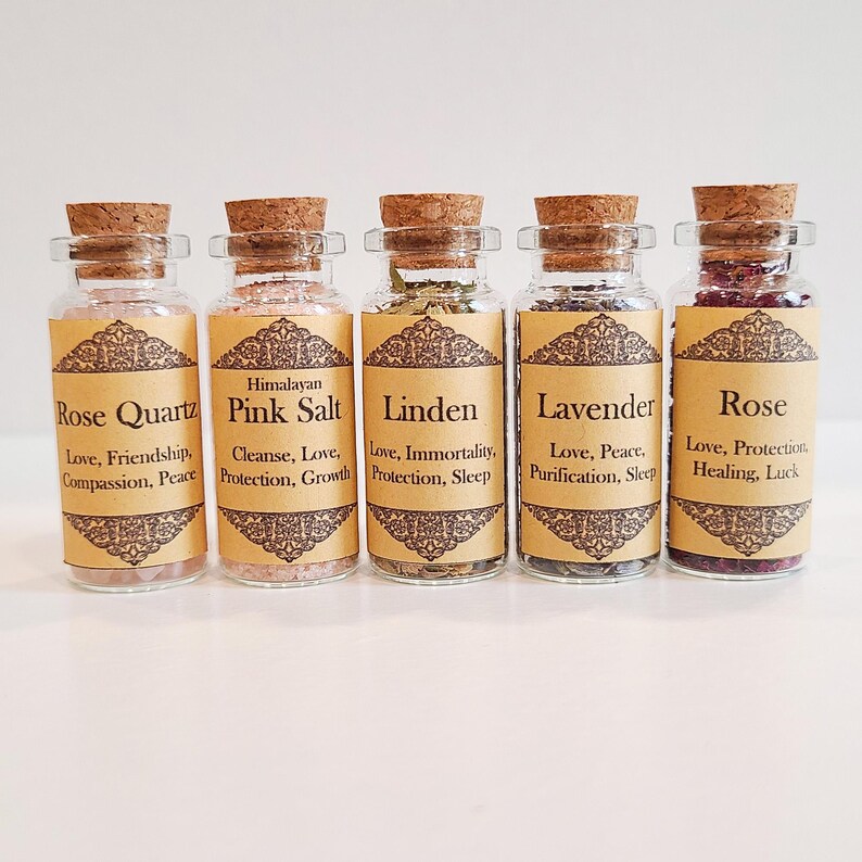 May include: Five small glass bottles with cork tops, each filled with different herbs and crystals. The bottles are labeled with the names of the herbs and crystals, including Rose Quartz, Himalayan Pink Salt, Linden, Lavender, and Rose. Each label also includes a description of the herb or crystal's properties, such as love, friendship, compassion, peace, cleanse, protection, growth, immortality, sleep, purification, healing, and luck.