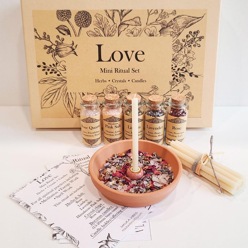 May include: A love ritual set with herbs, crystals, and candles in a brown box. The set includes rose quartz, pink salt, linden, lavender, and rose. The set also includes a small terracotta bowl with dried herbs and crystals, a white candle, and a bundle of white taper candles.