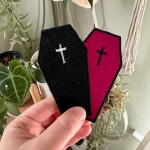 May include: Two felt coffin-shaped patches with embroidered crosses. The patch on the left is black with a white cross, and the patch on the right is red with a black cross.