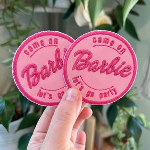May include: Two pink round iron-on patches with white stitching. The patches have the text "Come on Barbie, let's go party" in a pink cursive font.