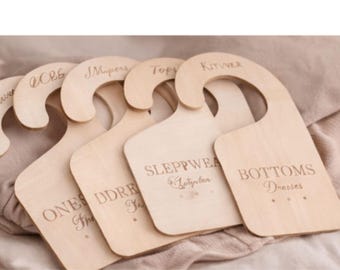 Puede incluir: Un conjunto de separadores de armario de madera con etiquetas grabadas. Las etiquetas incluyen "Musers", "Ones", "Sleepwear" y "Bottoms". Los separadores son de color claro y tienen una parte superior curvada para colgar.