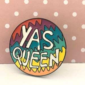 May include: A circular badge featuring the phrase "YAS QUEEN" in white lettering, outlined in black. The background is a vibrant, wavy pattern in purple, teal, yellow, and orange. The badge is edged with a black border.