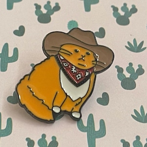 May include: An orange cat enamel pin, sporting a brown cowboy hat and a red bandana with white and black patterns. The cat has a white chest and paws. The background is light pink, decorated with cactus and heart shapes.