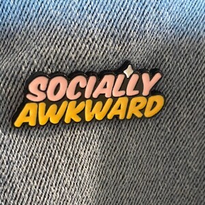 May include: A pin with the words "SOCIALLY AWKWARD" in a bubble letter style. The text is in pink and yellow with a black outline and a small star. The pin is on a denim background.