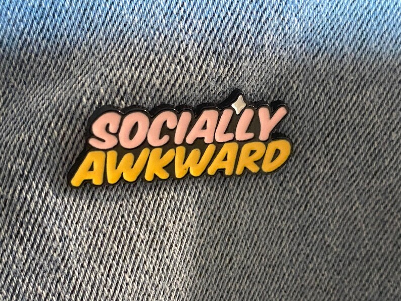 May include: A pin with the words "SOCIALLY AWKWARD" in a bubble letter style. The text is in pink and yellow with a black outline and a small star. The pin is on a denim background.