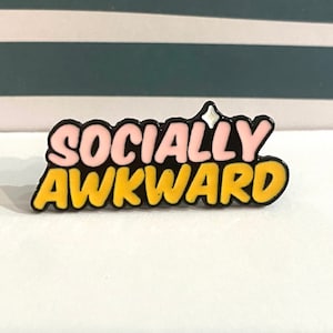 May include: A pin with the words "SOCIALLY AWKWARD" in a bubble letter style. The top letters are pink, and the bottom letters are yellow. The pin has a black border and a white accent at the top.