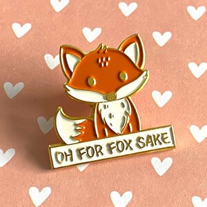 May include: A gold-edged enamel pin depicting a cartoon fox with orange and white fur. The fox holds a white sign with the text "OH FOR FOX SAKE". The pin is set against a pink background with white heart shapes. A humorous accessory.