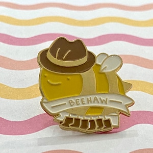 May include: A yellow and brown enamel pin of a cartoon bee wearing a brown fedora. The bee has white wings and a banner that reads "BEEHAW". The pin is set against a wavy pink, yellow, and white background.
