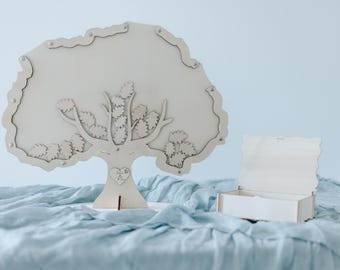 May include: A white wooden tree with leaves cut out, standing on a light blue fabric surface. A small white wooden box with a lid is also on the surface. The tree has a heart-shaped cutout at the base with the words "Love You" written inside.