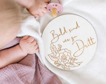 May include: A white wooden round sign with gold lettering that reads "Bald sind wir zu Dritt" with a floral design. The sign is on a white blanket with a baby's feet and a pink dummy.