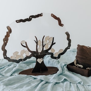 Könnte beinhalten: Eine hölzerne Alternative zum Gästebuch in Form eines Baumes, mit einer transparenten Acrylmitte und kleinen Blattausschnitten aus Holz. Daneben steht eine passende Holzkiste. Der Baum ist dunkelbraun mit einem Herz-Ausschnitt.