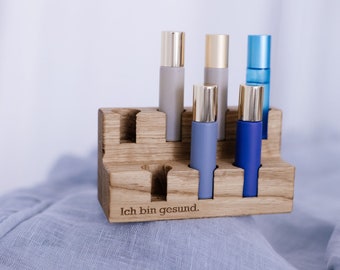 May include: Wooden stand with six slots for essential oil bottles. The stand is engraved with the text "Ich bin gesund."  The bottles are various shades of blue and gray.
