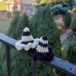 Puede incluir: Dos sombreros de bruja de ganchillo. Un sombrero es de rayas blancas y negras con un borde color crema. El otro sombrero es de rayas blancas y negras. Los sombreros están sobre una barandilla de metal negro con un fondo verde borroso.