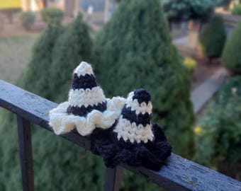 May include: Two crocheted witch hats. One hat is black and white striped with a cream-colored brim. The other hat is black and white striped. The hats are sitting on a black metal railing with a blurred green background.