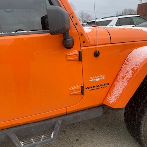 Puede incluir: Jeep Wrangler Unlimited Sahara naranja con un escalón lateral negro y nieve en la carrocería. El Jeep tiene un espejo lateral negro y una antena negra.