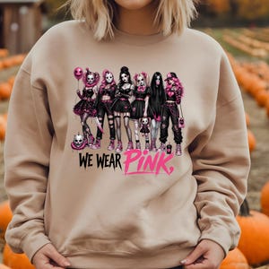 A person wearing a beige sweatshirt with a graphic design featuring a group of women in dark clothing and makeup, along with the text "WE WEAR PINK" printed on the sweatshirt.
