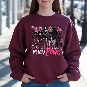 A woman wearing a burgundy sweatshirt with a graphic design featuring a group of women in dark clothing and makeup, along with the text "WE WEAR PINK."