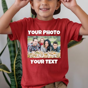 May include: A red short-sleeved t-shirt featuring a rectangular photo print of a family sitting in autumn leaves. The text "YOUR PHOTO" is printed in white above the image, and the text "YOUR TEXT" is printed in white below the image.