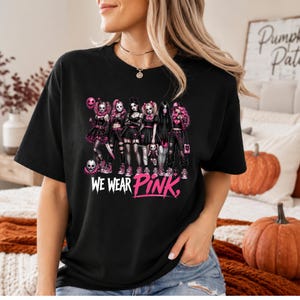 A woman wearing a black t-shirt with a graphic design featuring a group of women in various outfits, including a skull and crossbones, and the text "WE WEAR PINK" printed on the shirt.