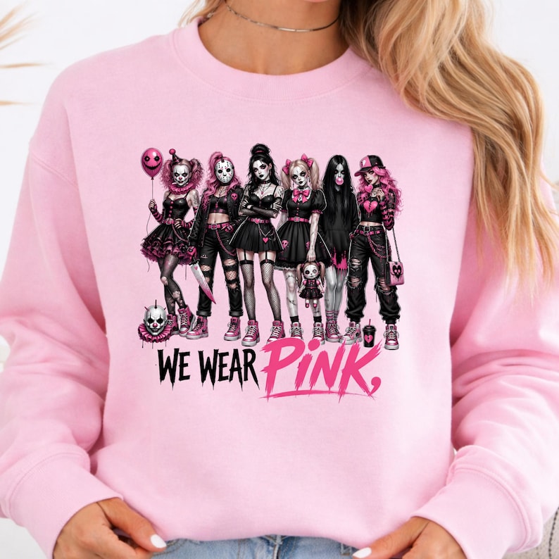 A woman wearing a pink sweatshirt with a graphic design featuring a group of women dressed in dark, gothic-style clothing and makeup, along with the text "WE WEAR PINK."