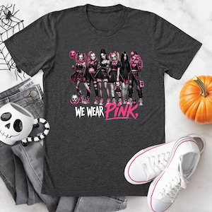 a black t-shirt with a graphic design of a group of women dressed in black and pink outfits, along with the text "WE WEAR PINK." The t-shirt is displayed on a white surface, accompanied by a pair of jeans, a pumpkin, and a pair of white sneakers.
