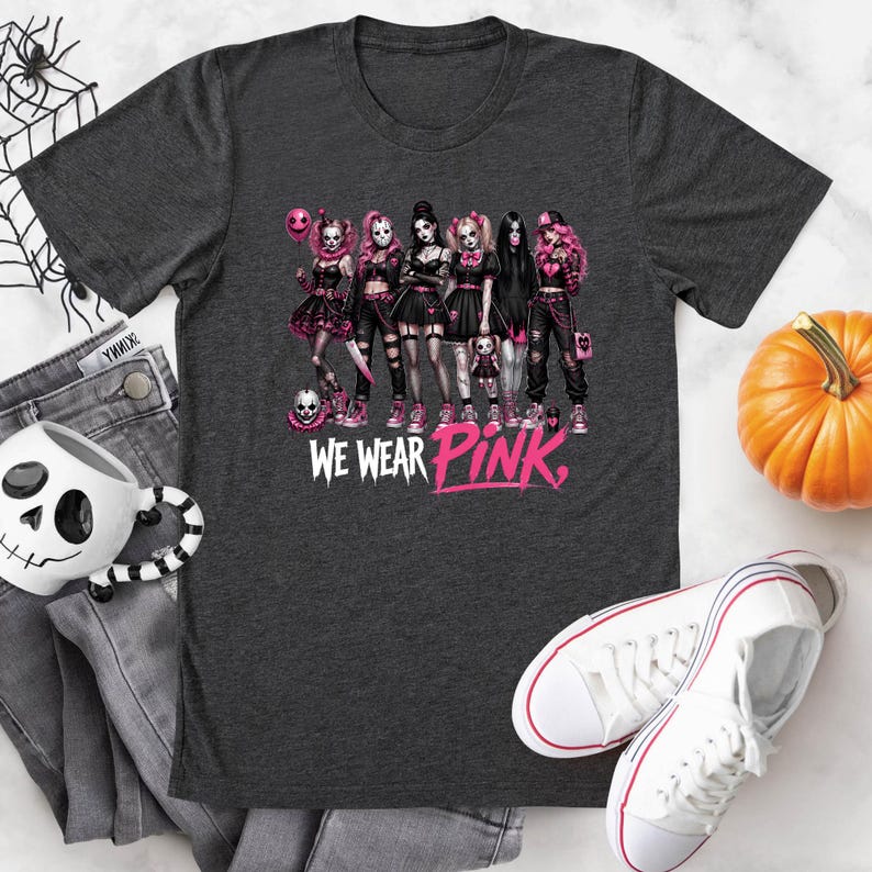 a black t-shirt with a graphic design of a group of women dressed in black and pink outfits, along with the text "WE WEAR PINK." The t-shirt is displayed on a white surface, accompanied by a pair of jeans, a pumpkin, and a pair of white sneakers.
