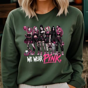 A woman wearing a green sweater with a graphic design featuring a group of women with various styles and accessories, including skull-like elements.