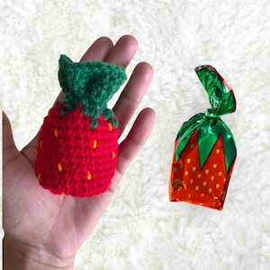 May include: A red crocheted strawberry-shaped pouch with green leaves and a red and green foil-wrapped candy with a strawberry design.