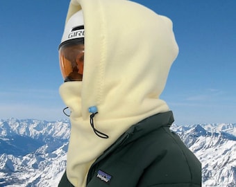 May include: A cream-colored fleece balaclava with a drawstring closure and a blue toggle. The balaclava is worn over a white ski helmet with orange goggles and a dark green jacket. The background features a mountain range.
